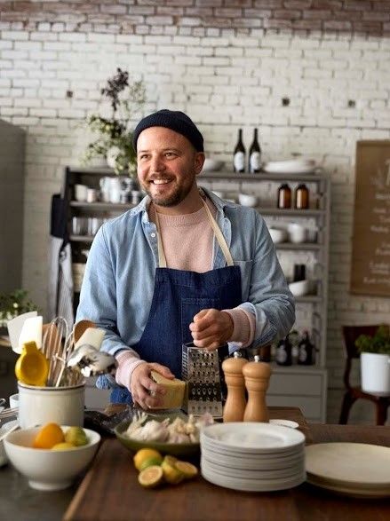 Chef Joshua McFadden in his element: the kitchen. The Portland-based chef is wearing a denim button down, a navy rolled up beanie, and is smiling while grating parmesan cheese.