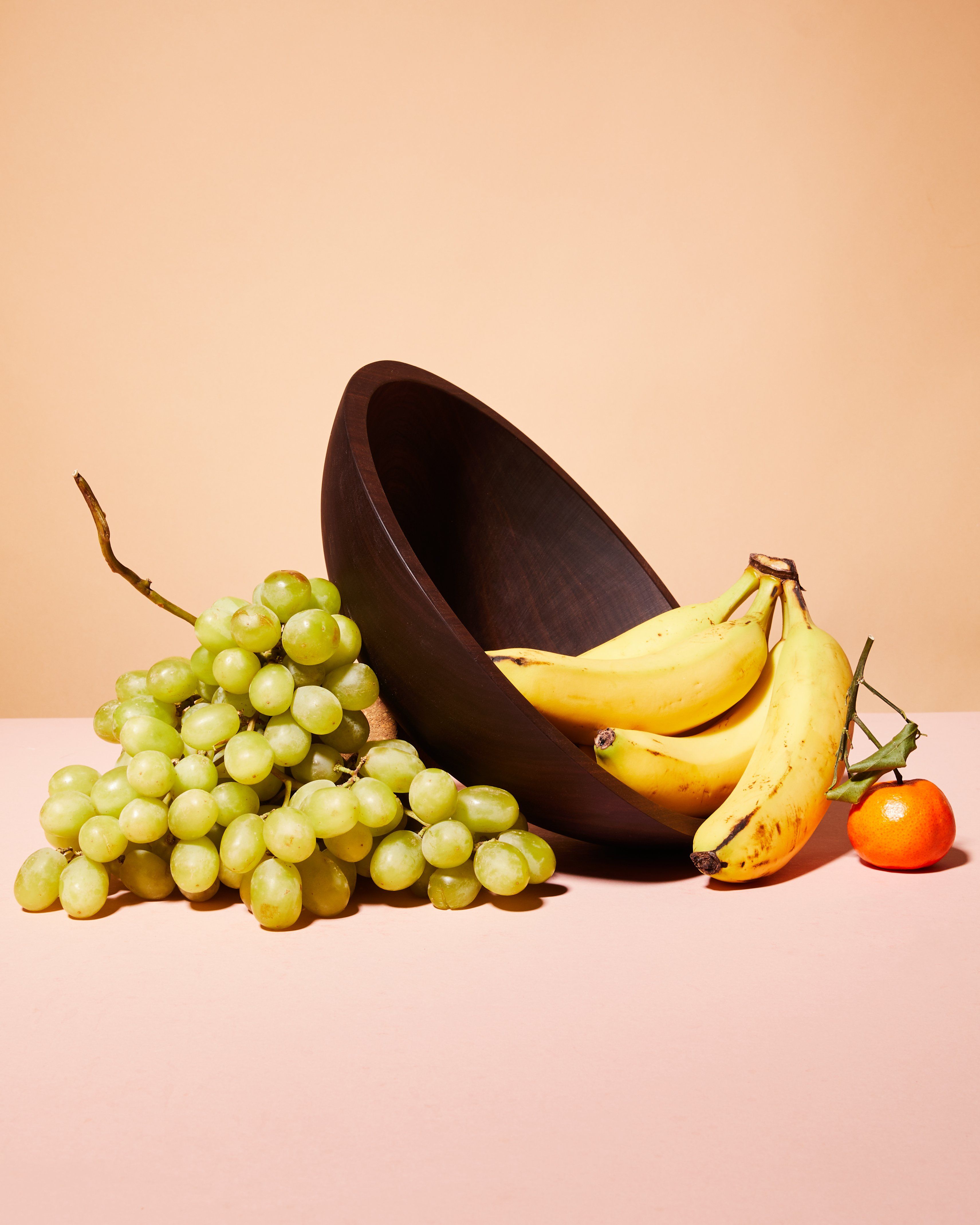 A large walnut wooden bowl is tilted artfully sitting on a bunch of grapes and has bananas and a persimmon spilling out on the other side