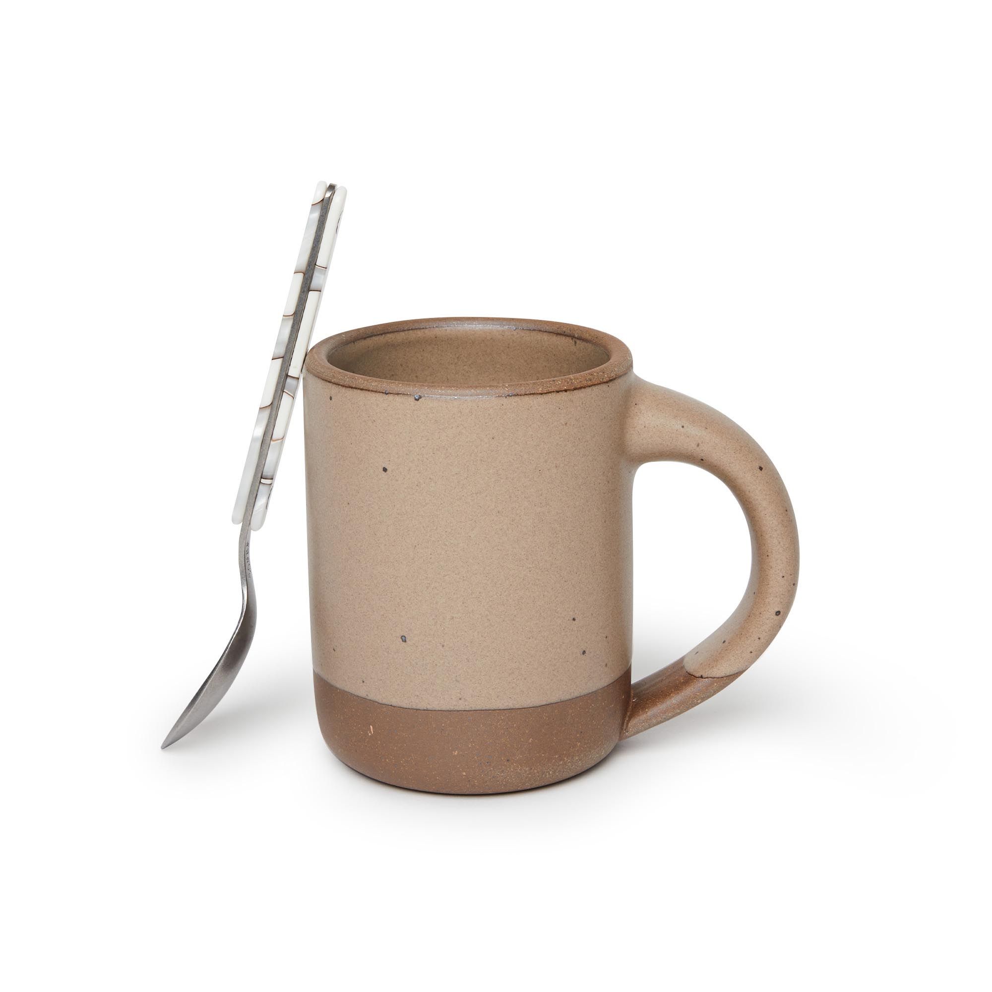 A spoon leaning on a medium sized ceramic mug with handle in a warm pale brown color featuring iron speckles and unglazed rim and bottom base.