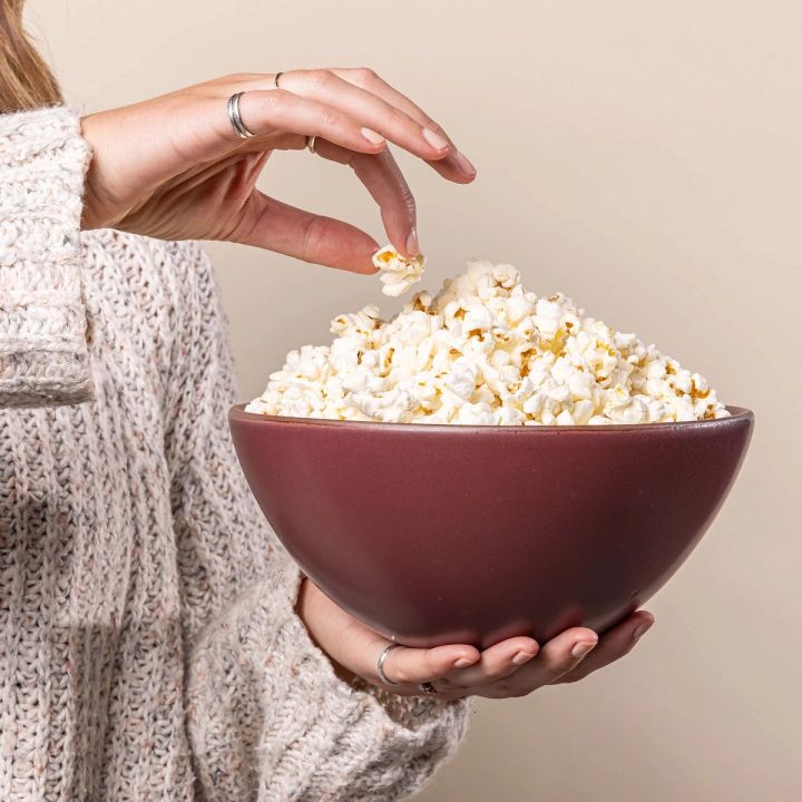 A hands holds a large rounded ceramic bowl in a plum color featuring an unglazed rim and is grabbing a piece of popcorn that fills the bowl.