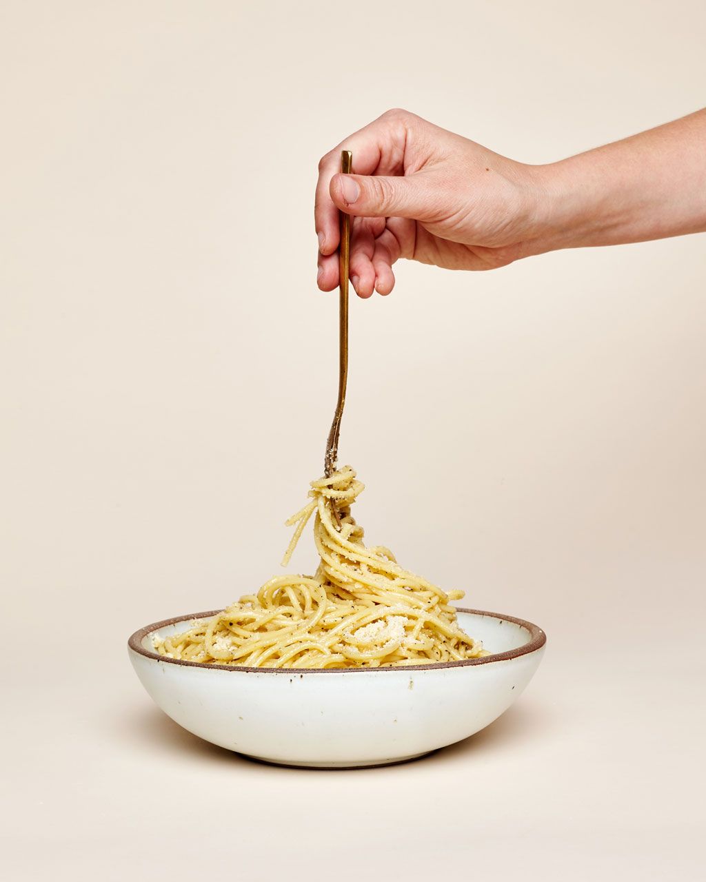 A hand is holding a fork twirling spaghetti in a shallow dinner bowl in a cool white color.
