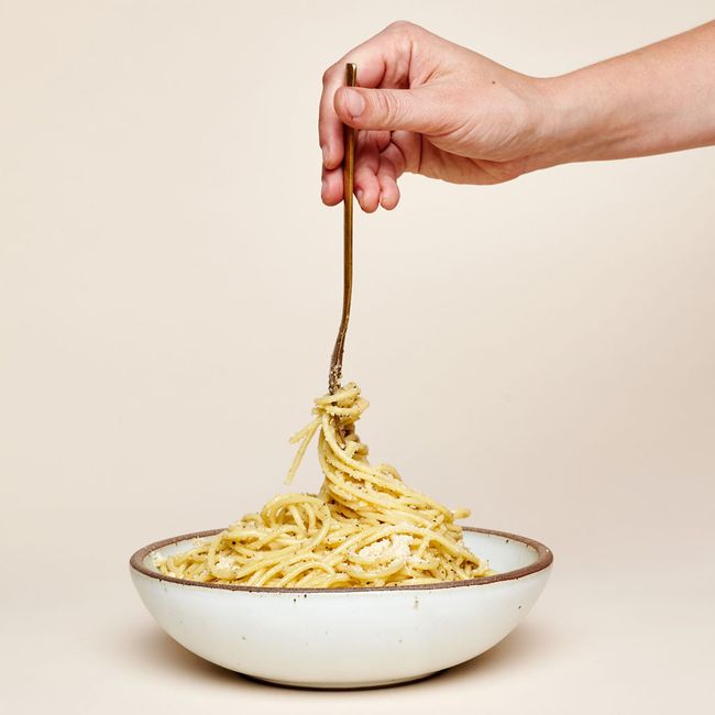 A hand is holding a fork twirling spaghetti in a shallow dinner bowl in a cool white color.