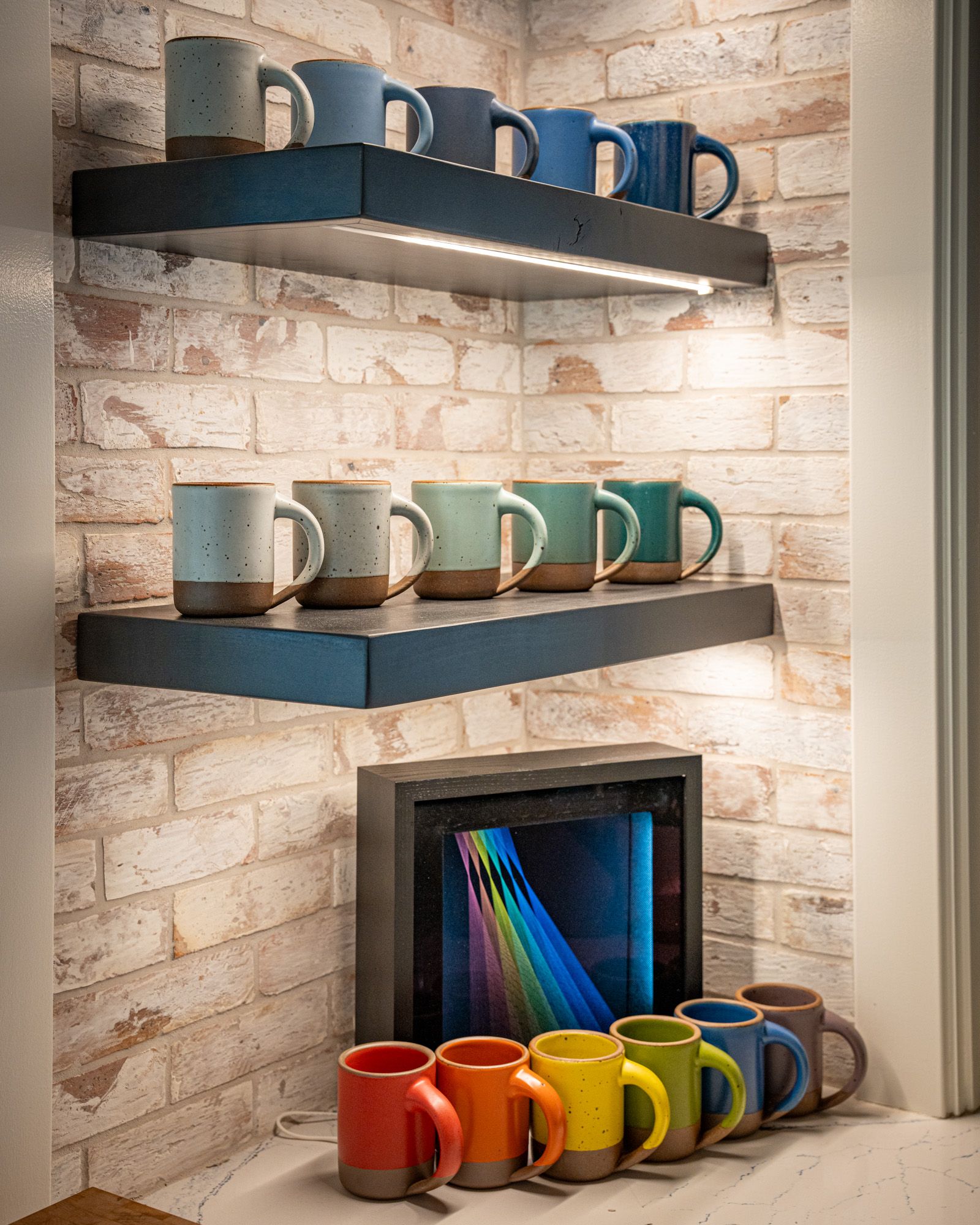 Cornered kitchen shelves are filled with ceramic mugs in different blue hues and a kitchen countertop has mugs in each color the rainbow.