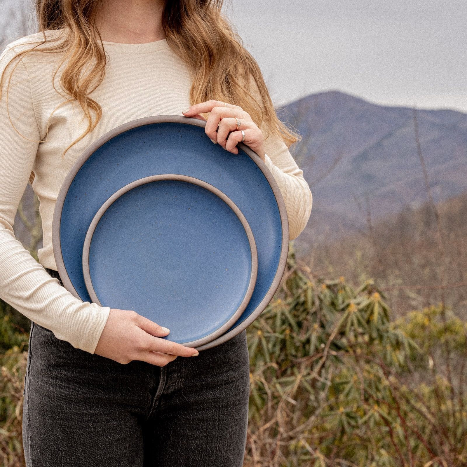 A woman outside holding a ceramic serving platter and plate in a cool medium blue, against a background of a mountain landscape