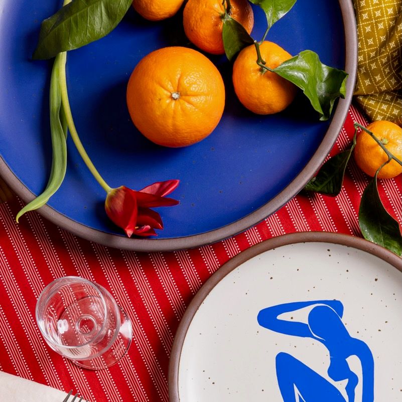 A closeup of a large ceramic platter in a cobalt blue color with oranges and flowers, and next to a plate with a blue decal of a nude figure.