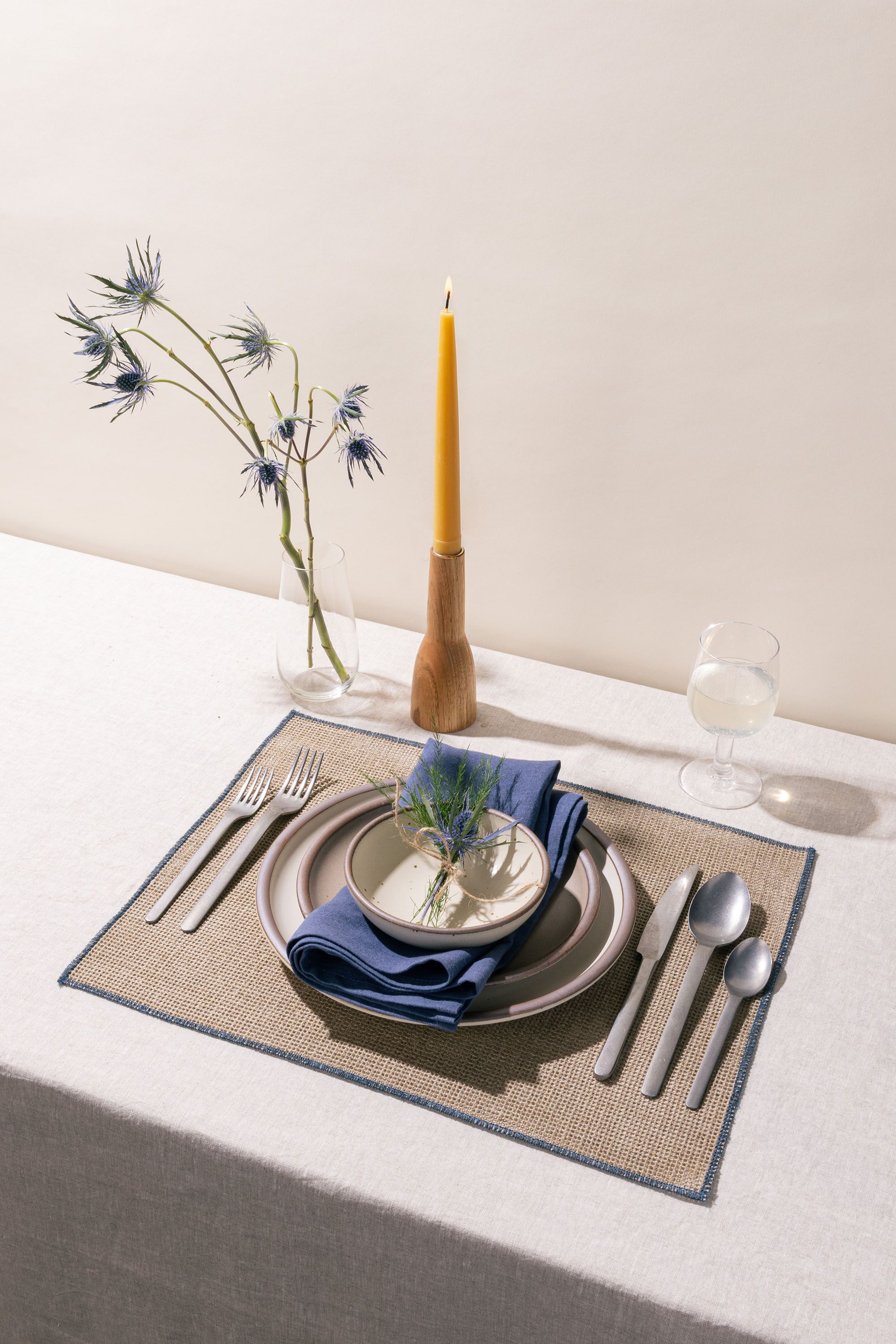 A table setting featuring ceramic plates in neutral colors, a blue napkin, a taper candle, and steel flatware.