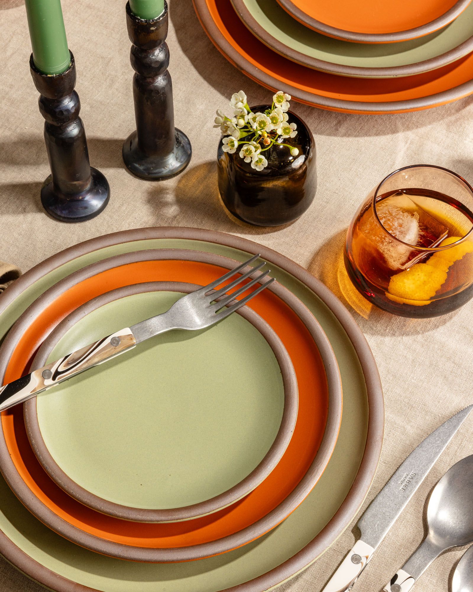 An overhead table setting featuring sage green and bold orange plates, sophisticated marbled flatware, candlesticks, and a whiskey snifter with a cocktail.