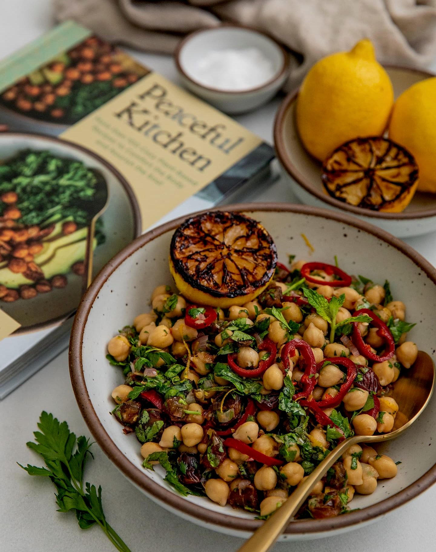 A plate filled with a chickpeas, vegetables, and lemon next to a cookbook titled, 'Peaceful Kitchen'