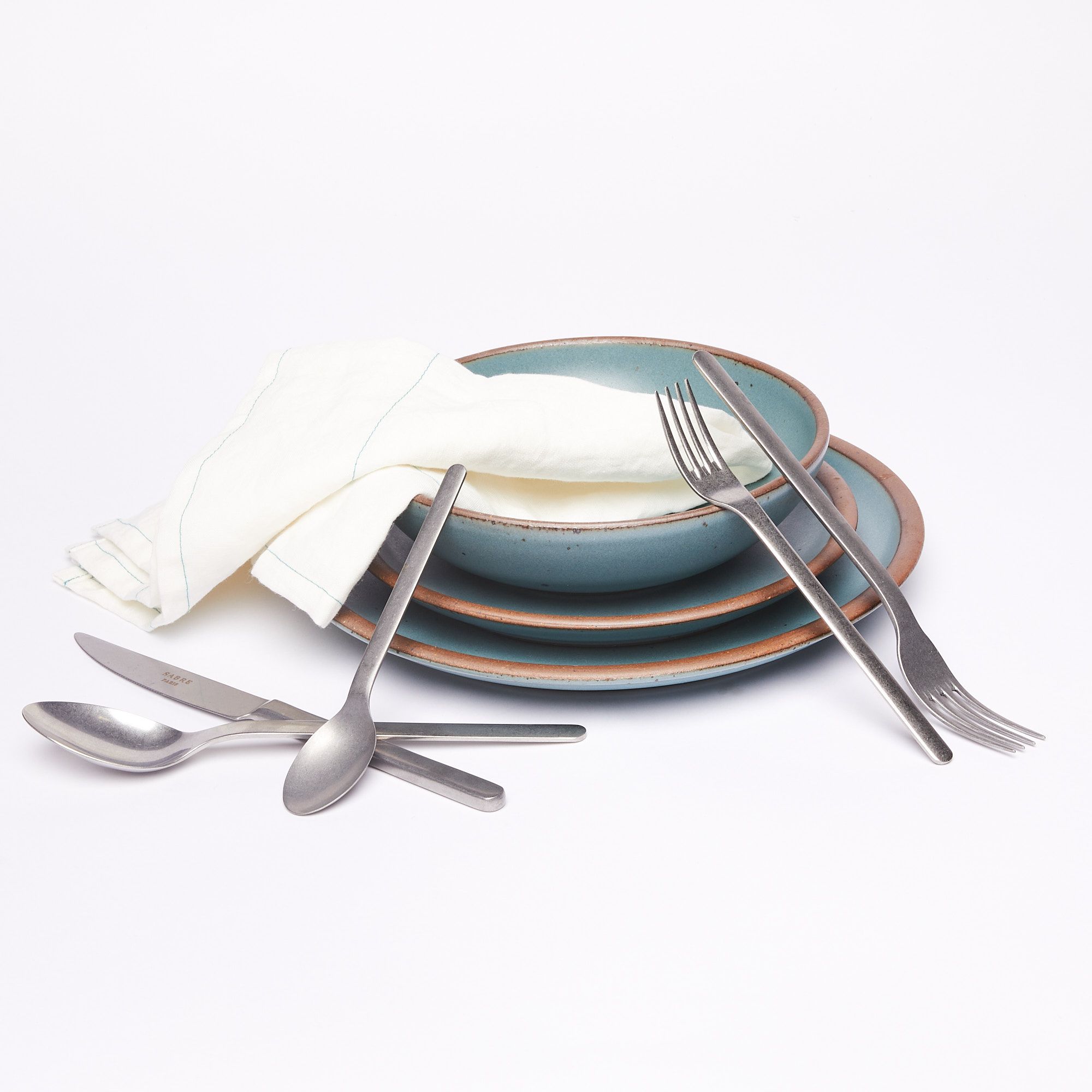 An everyday bowl, side plate, and dinner plate in turquoise stacked with a linen napkin surround by five pieces of stainless steel flatware