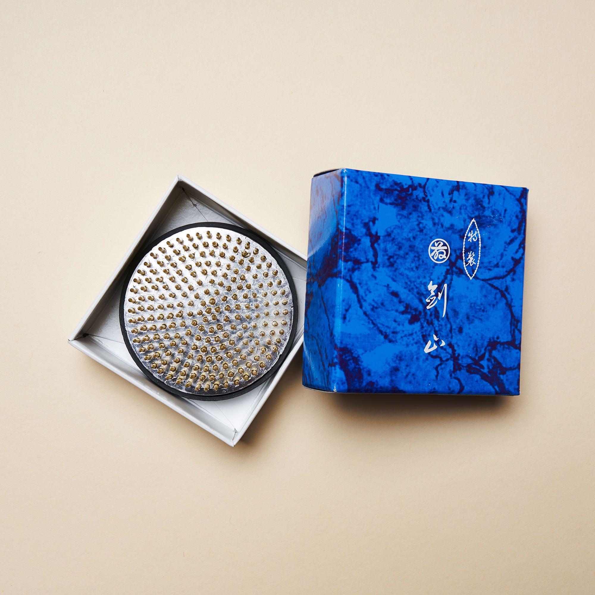 A medium "kenzan" – a pin frog made of brass spikes on a flat circular base to assist in flower arrangements sitting inside a white box with a blue top on the right