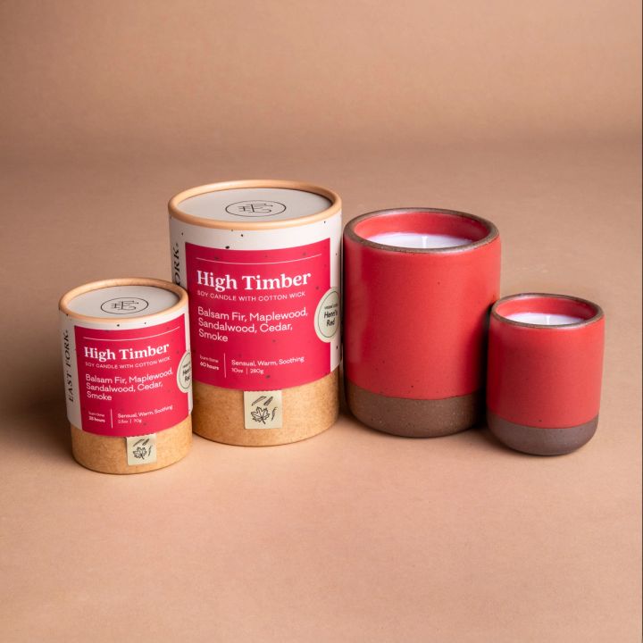 Small and large ceramic vessel next to each other in bold red color with candles inside each. Cardboard tube packaging is on the left with branding stickers that say "High Timber".