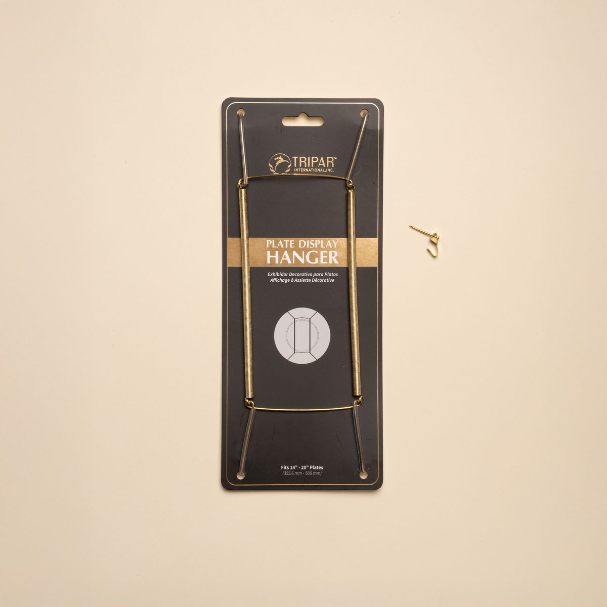 A brass plate hanger in its black cardboard package, with a nail off to the side.