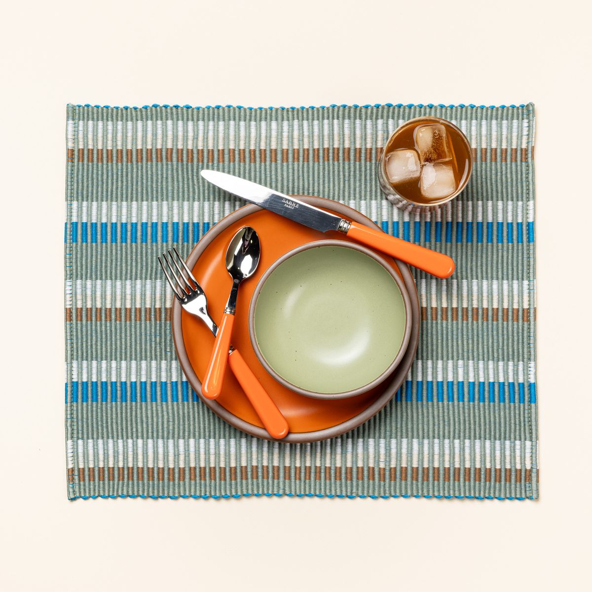 An orange and sage green place setting sits on a rectangle placemat with offset stripe design that is hand woven in sage, blue, and orange colors. Also on the placemat are a cocktail in a clear glass and a bold orange handled fork, spoon, and knife.