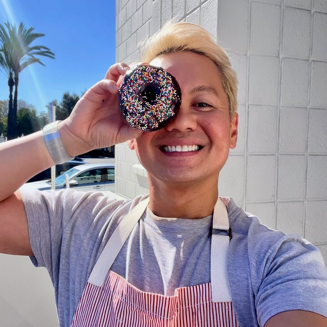 Ish is outside smiling wearing an apron and holding a chocolate sprinkled donut in one hand and holding it over his right eye.