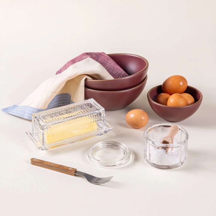 A group of kitchen items such as a bowl of eggs, butter dish, salt cellar fork, and a napkin