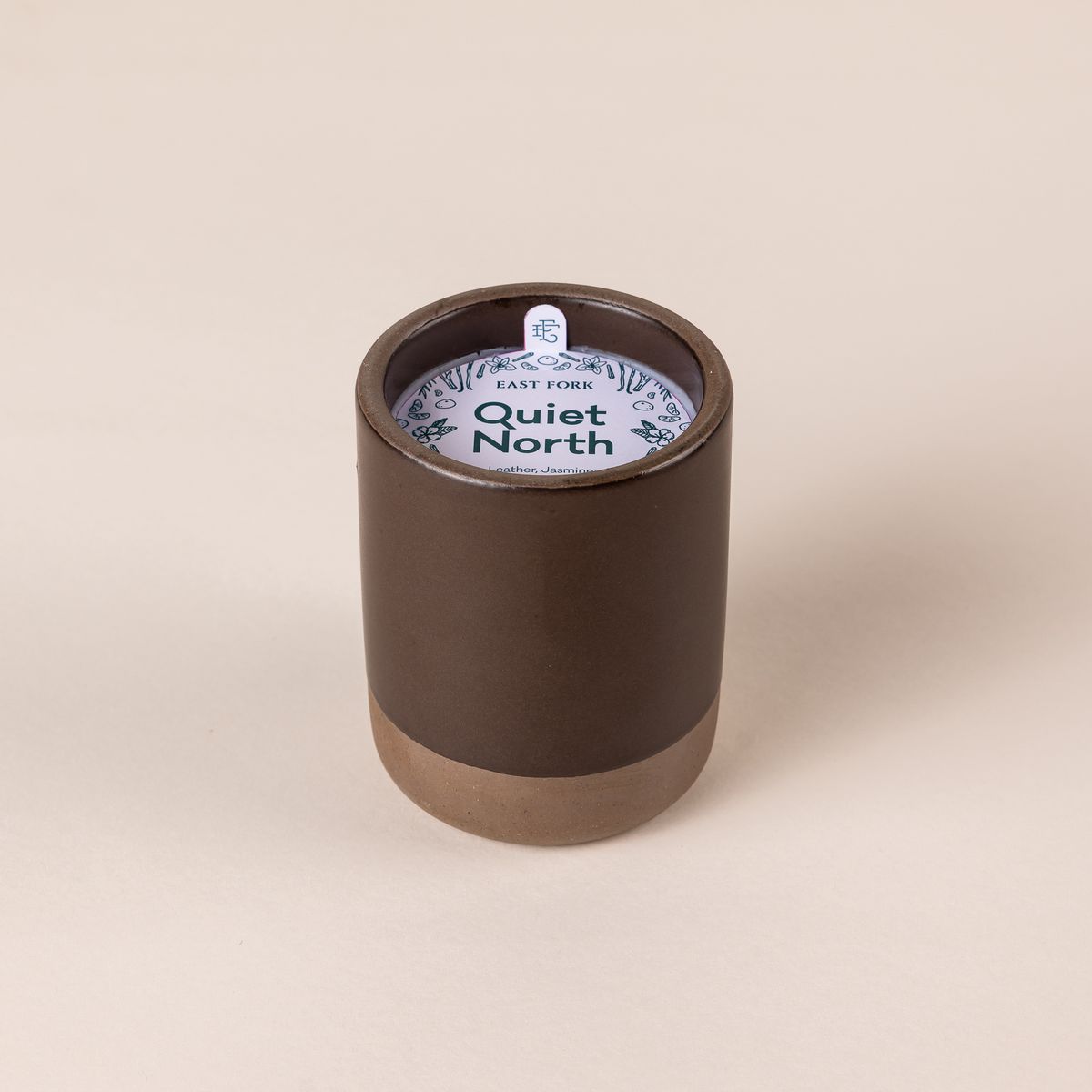 Large ceramic vessel in a dark cool brown color with candle inside. On top is a packaging label sitting on top that reads "Quiet North"