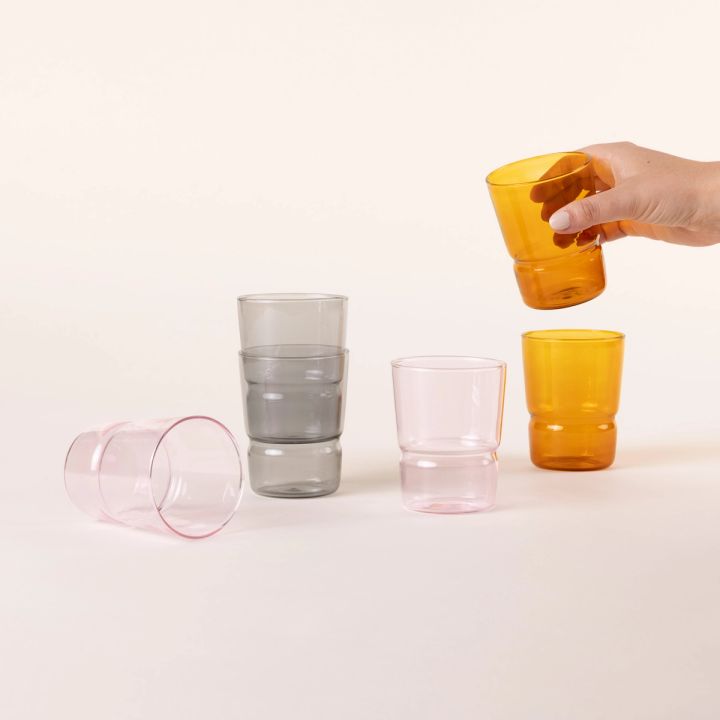 Several tumbler glasses that widen at the top in a grey, pink, and amber colors with a hand holding one.