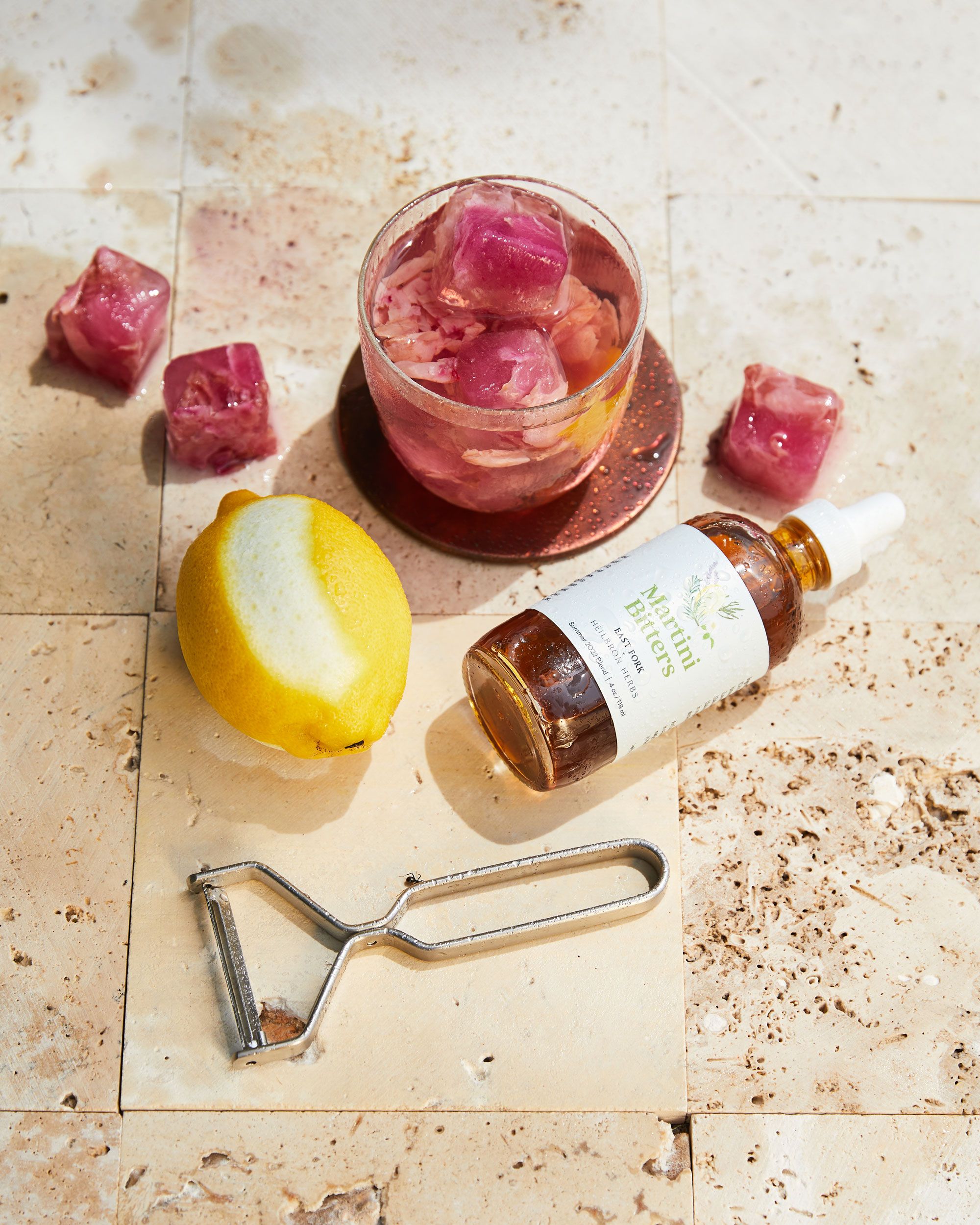 Rose petal ice cubes, lemon with a peel taken out, peeler, bitters bottle and short clear glass with cocktail in it