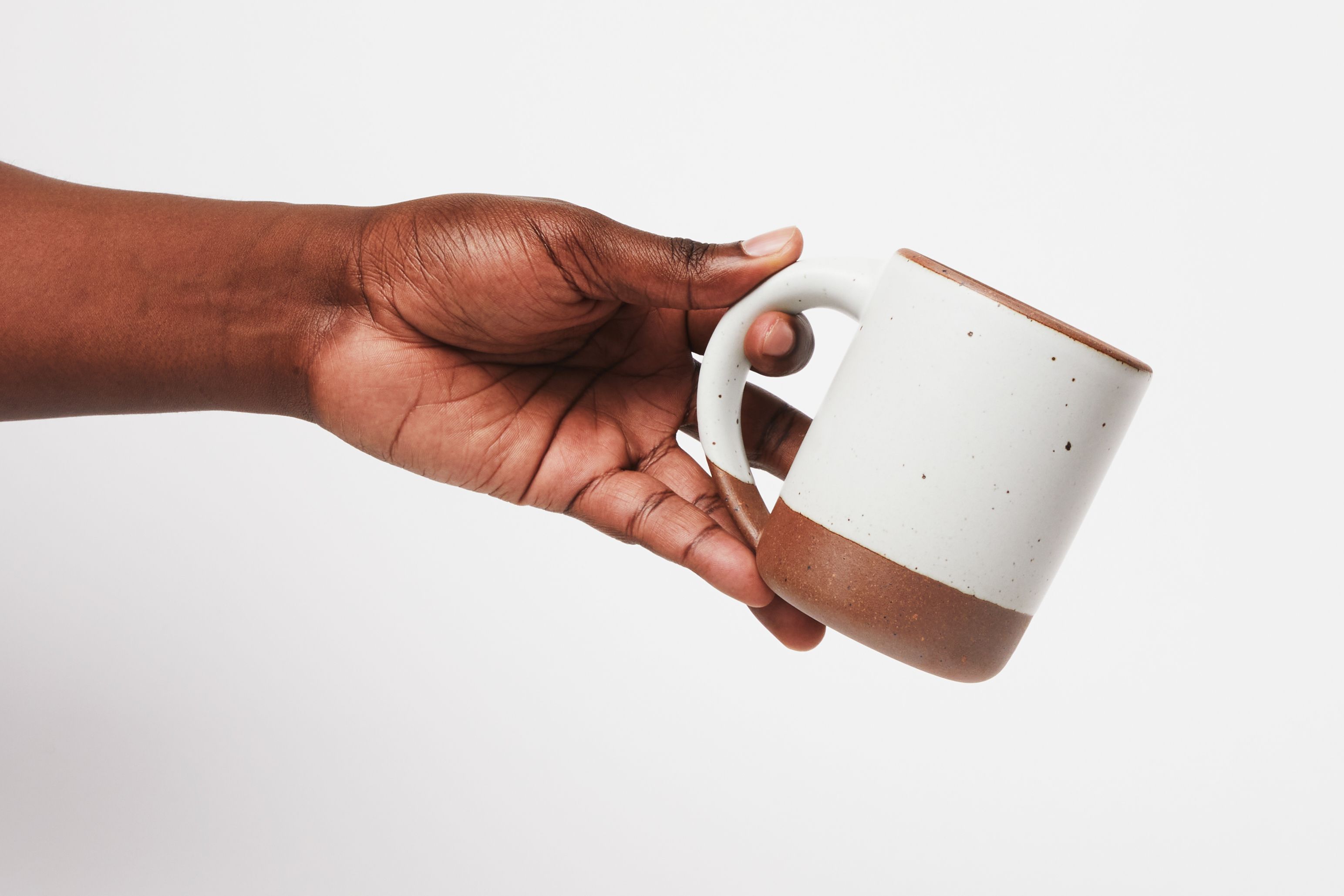Hand holding The East Fork Mug, a vessel with a handle, in a white glaze with unglazed rim and base.