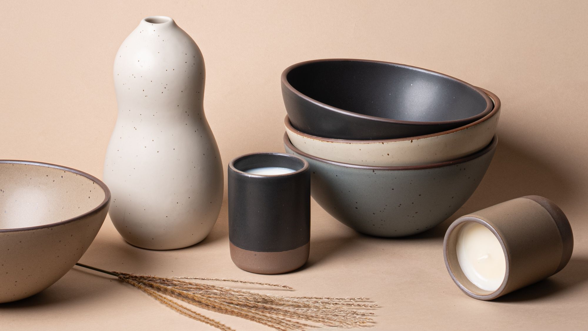 A neutral-toned arrangement of East Fork pottery, including bowls, a gourd-shaped vase, and ceramic candles, styled against a warm beige backdrop with dried grass accents.