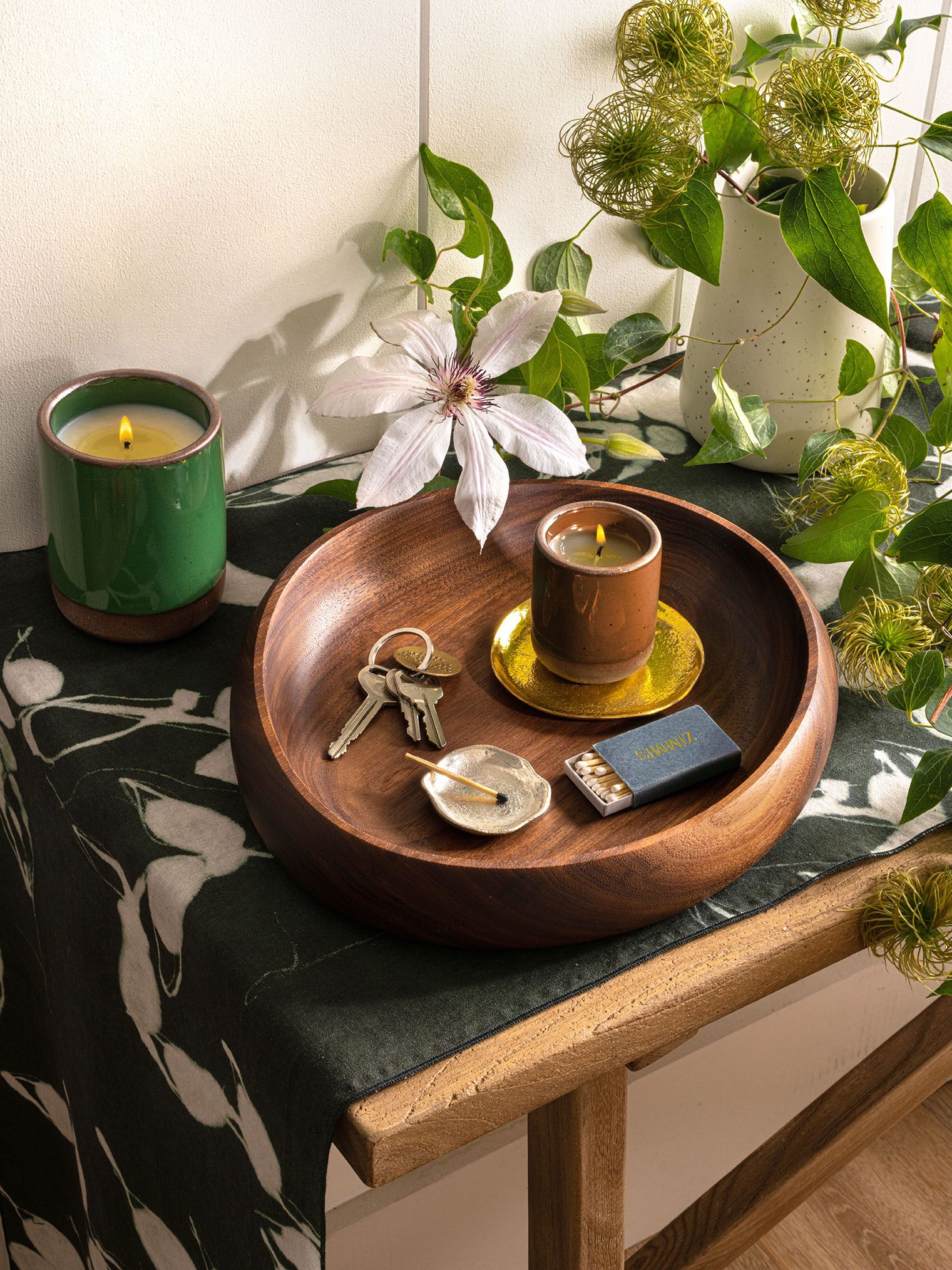 An entry table with a wooden tray with a candle, keys, and matches beside a blooming clematis and lush greenery.
