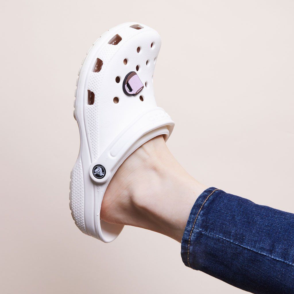 A denim-clad leg, bare ankle and white plastic clog on the foot. Clog has a charm that looks like an East Fork Mug