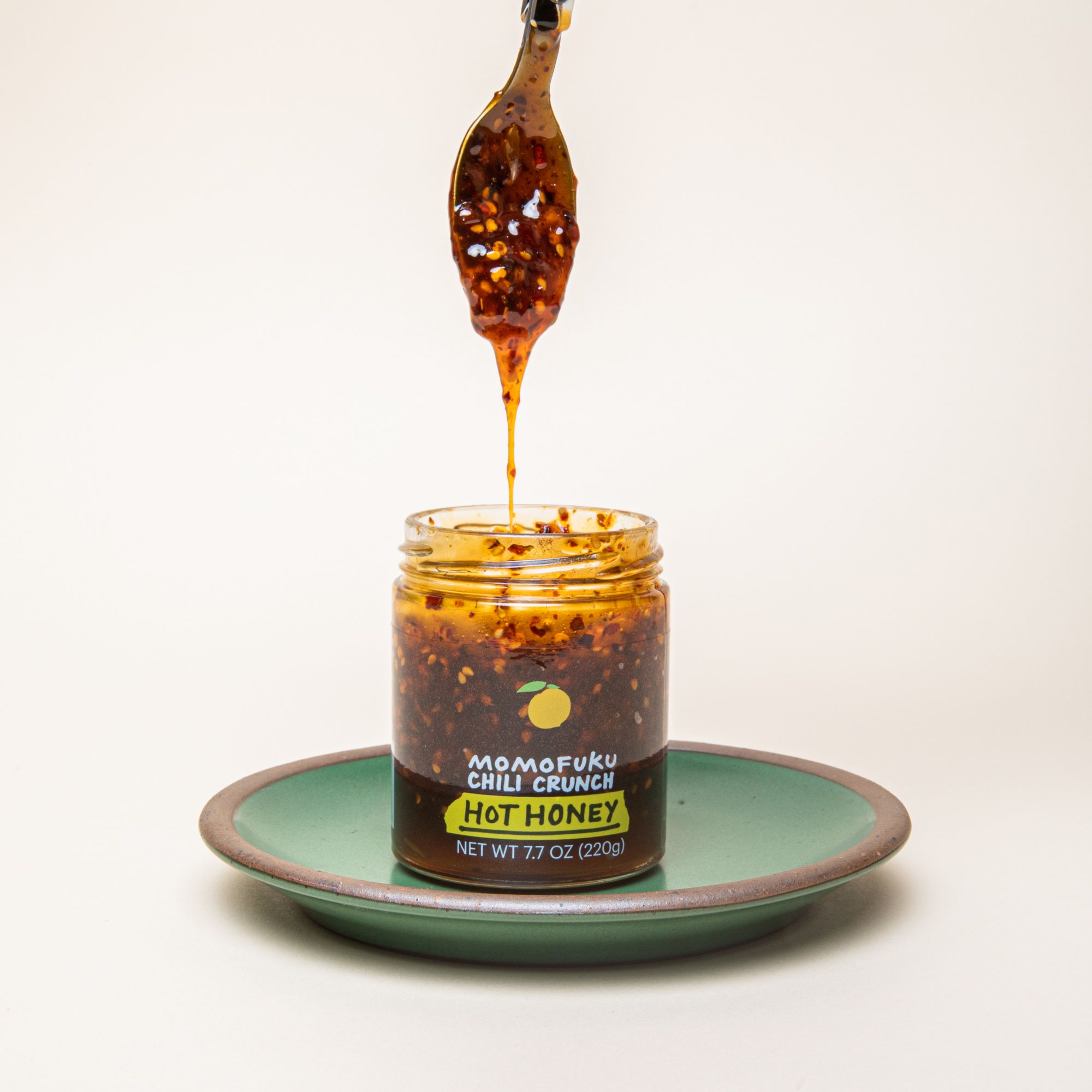 A spoon dripping some warm brown honey with chili flakes above a clear jar - the jar has a label that reads, "Momofuku Chili Crunch Hot Honey"