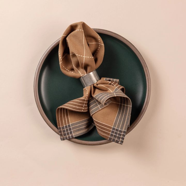 A folded light brown colored napkin pulled through a pewter napkin ring sits on a deep teal plate. The napkin is designed with cream gridlines with soft blue and brown striped edging.