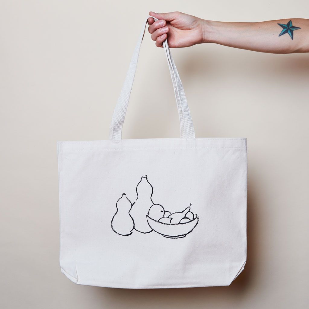 Extended arm with hand holding East Fork tote bag featuring pottery and fruit