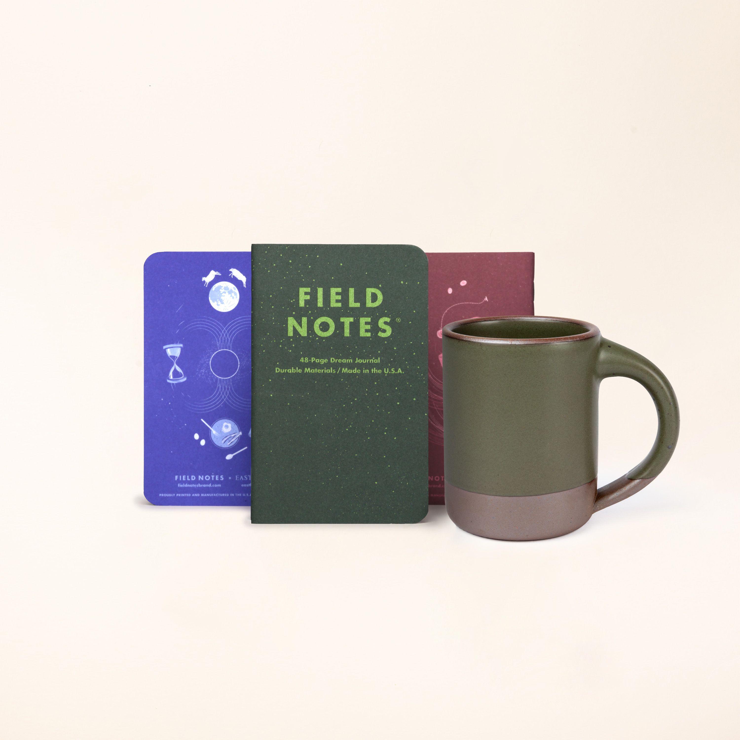 A 3 pack of Field Notes Journal standing up in a row in forest green, lapis blue, and plum colors with The Mug in a muted forest green color.