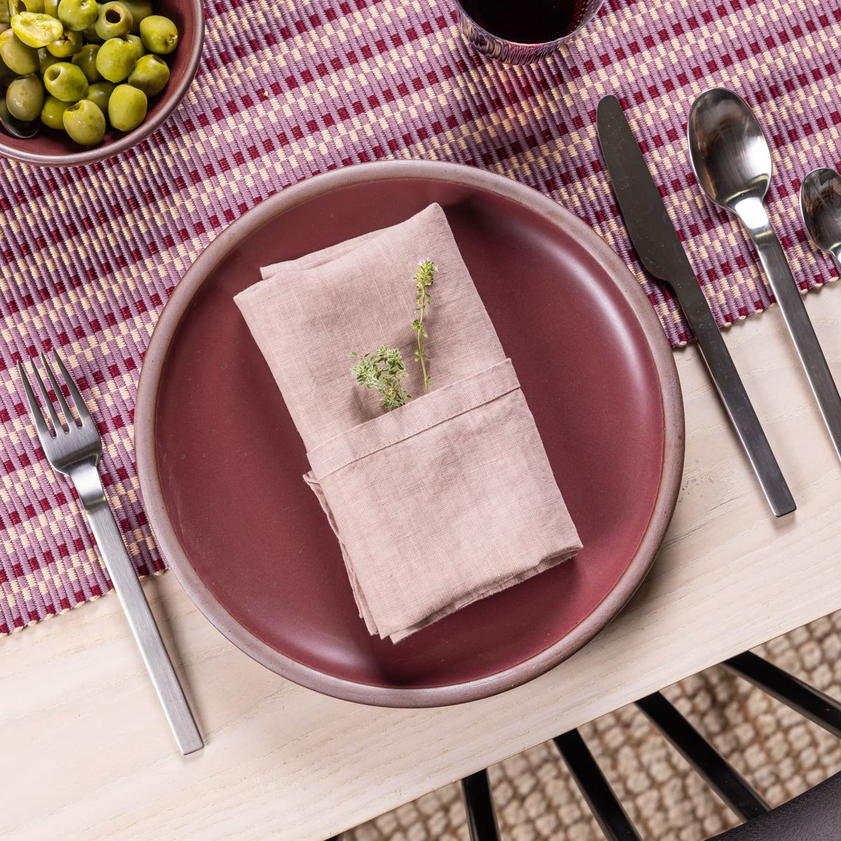 A folded linen napkin in a soft pink color sits with greenery on a plum ceramic plate with silverware