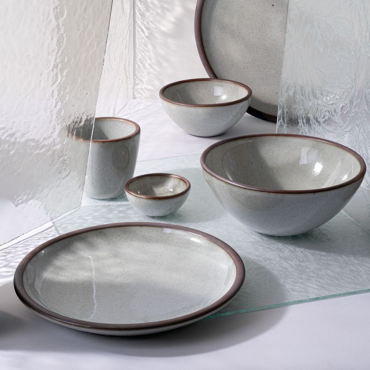 Set of ceramic dinnerware in a reflective icy white glaze with unglazed rims, arranged among textured glass panels on a white surface.