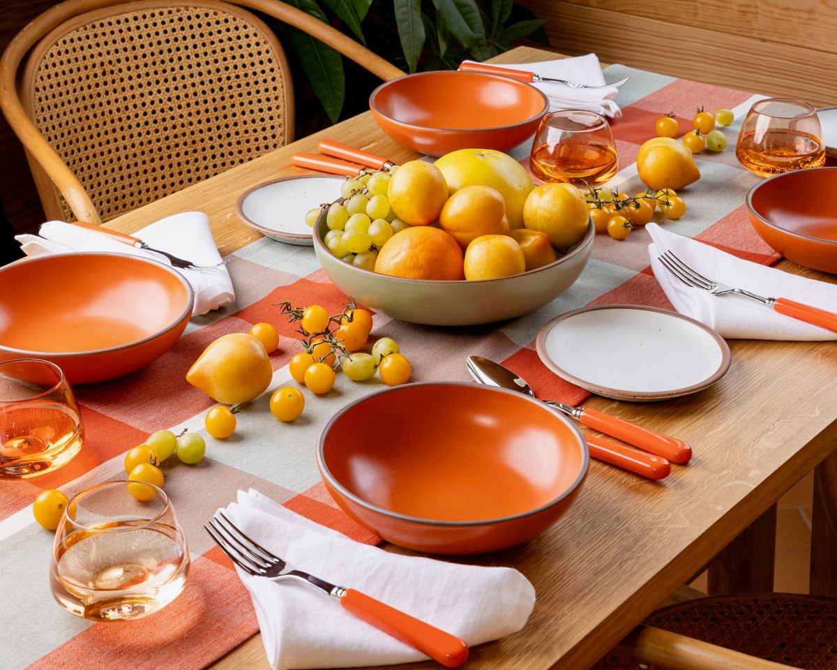 A table set with a table runner, bold orange bowls, orange-handled flatware, whiskey snifters, and a centerpiece of fruits and vegetables. Also pictured is a woven chair at the table.