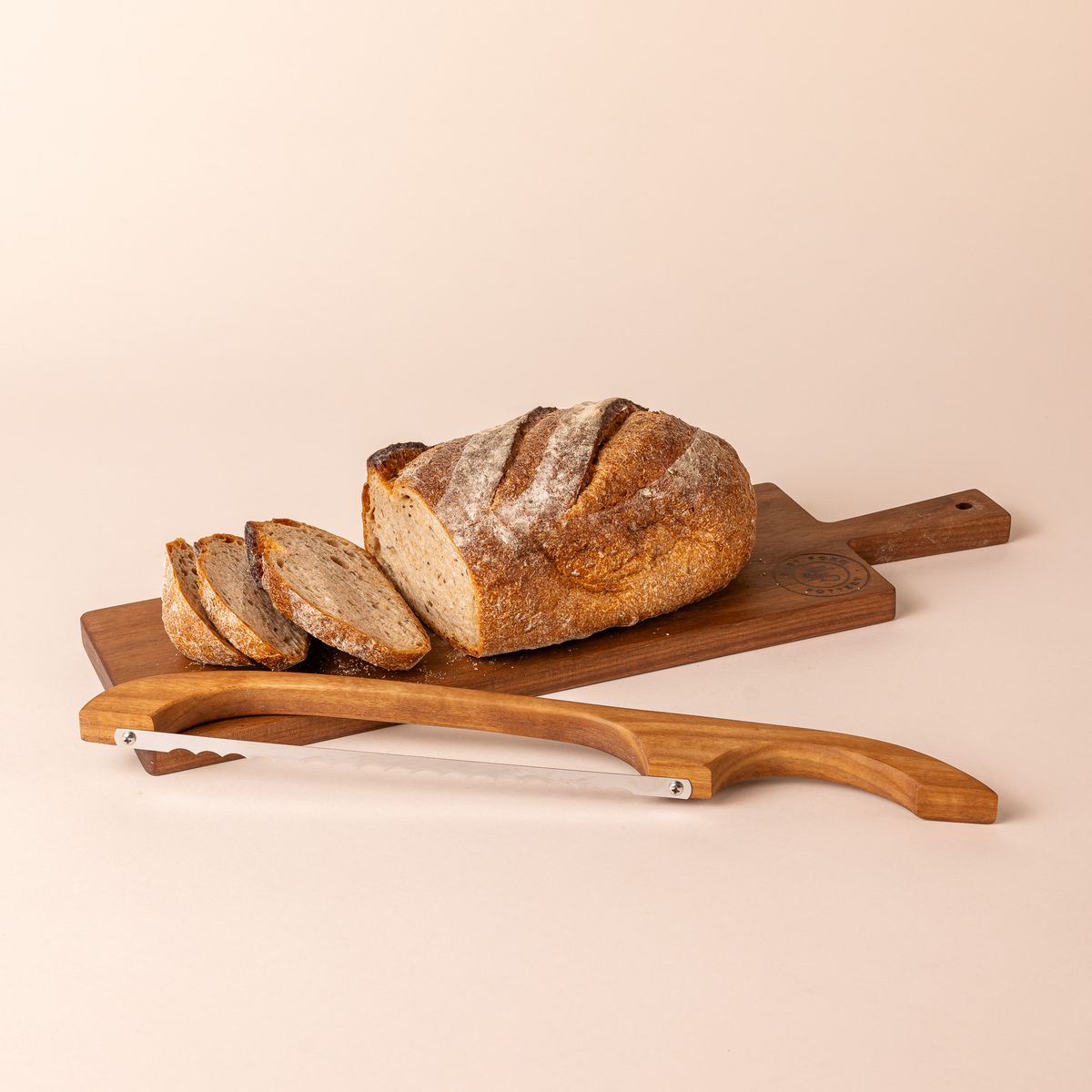 A loaf of bread with a few cut slices sits on a wood rectangle bread board with a handle. A matching wood bread bow sits on the side.
