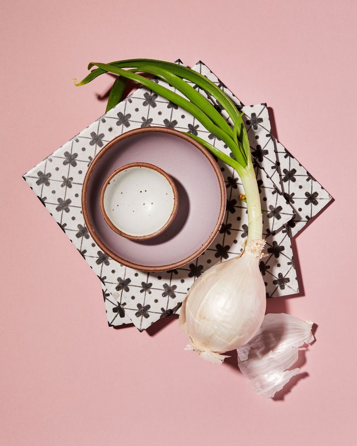 Two bowls, nestled inside one another. One light purple. The other bright white. Next to it, a large white onion, with the sprigs still intact, like you'd find at the farmers market. Two checkered tiles underneath the bowls and vegetable help frame the photo.