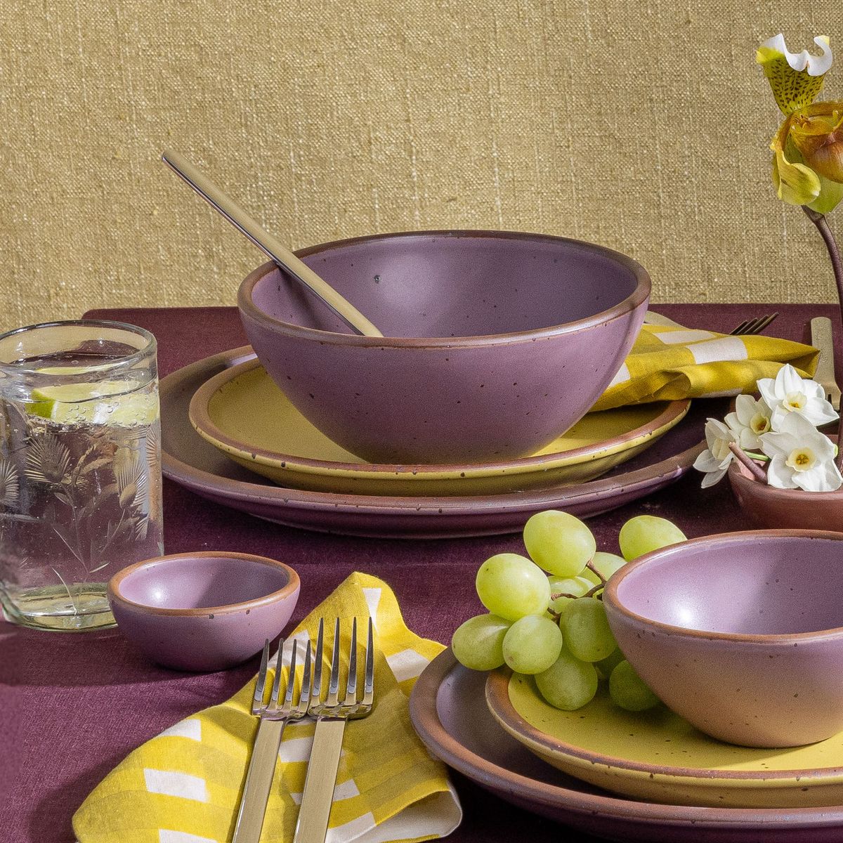 A tablescape featuring ceramic dinnerware in soft mauve purple bowls, and zesty yellow plates, as well as clear etched glassware, purple and yellow table linens, and silverware.