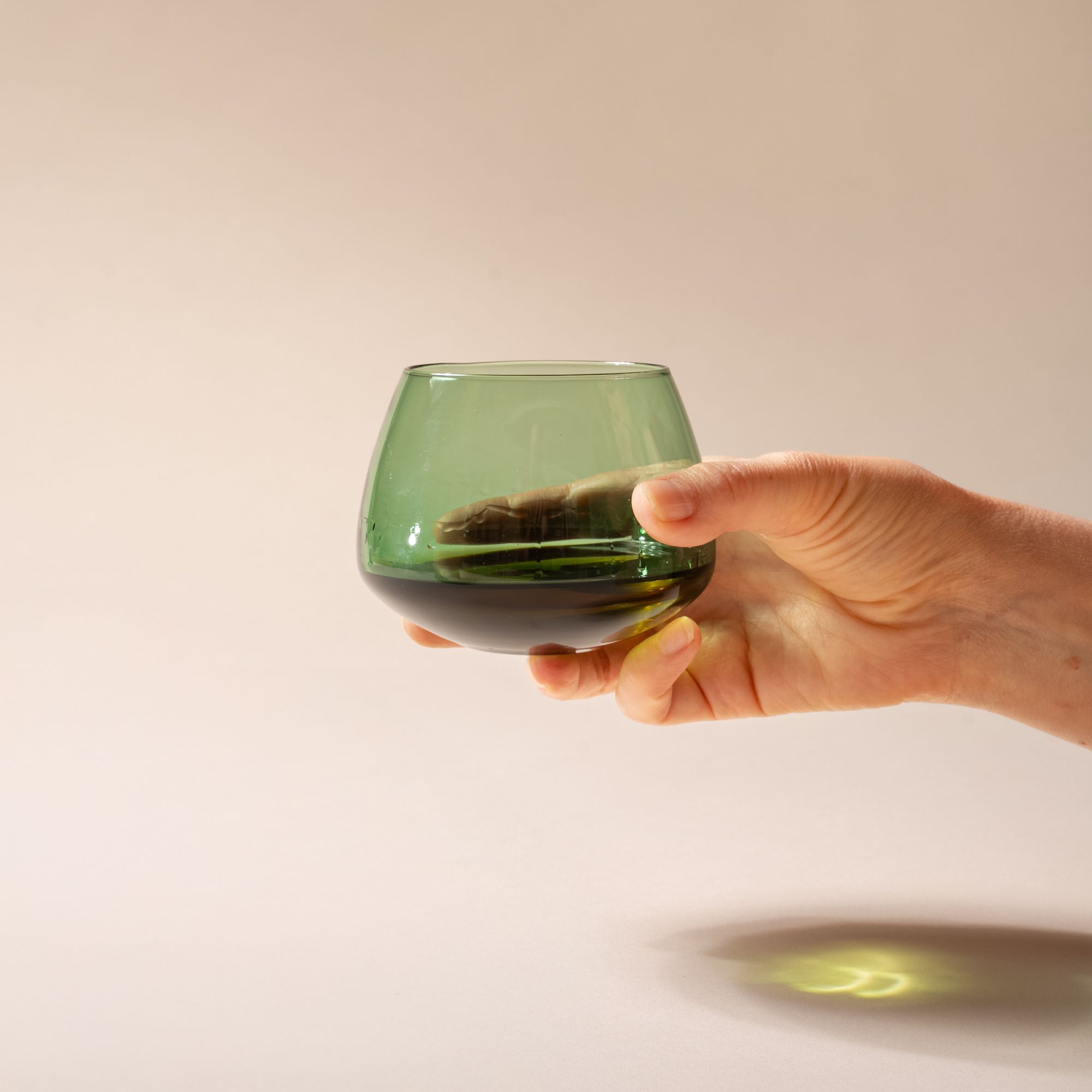 A hand holds a green glass whiskey snifter with a rounded base, with whiskey inside