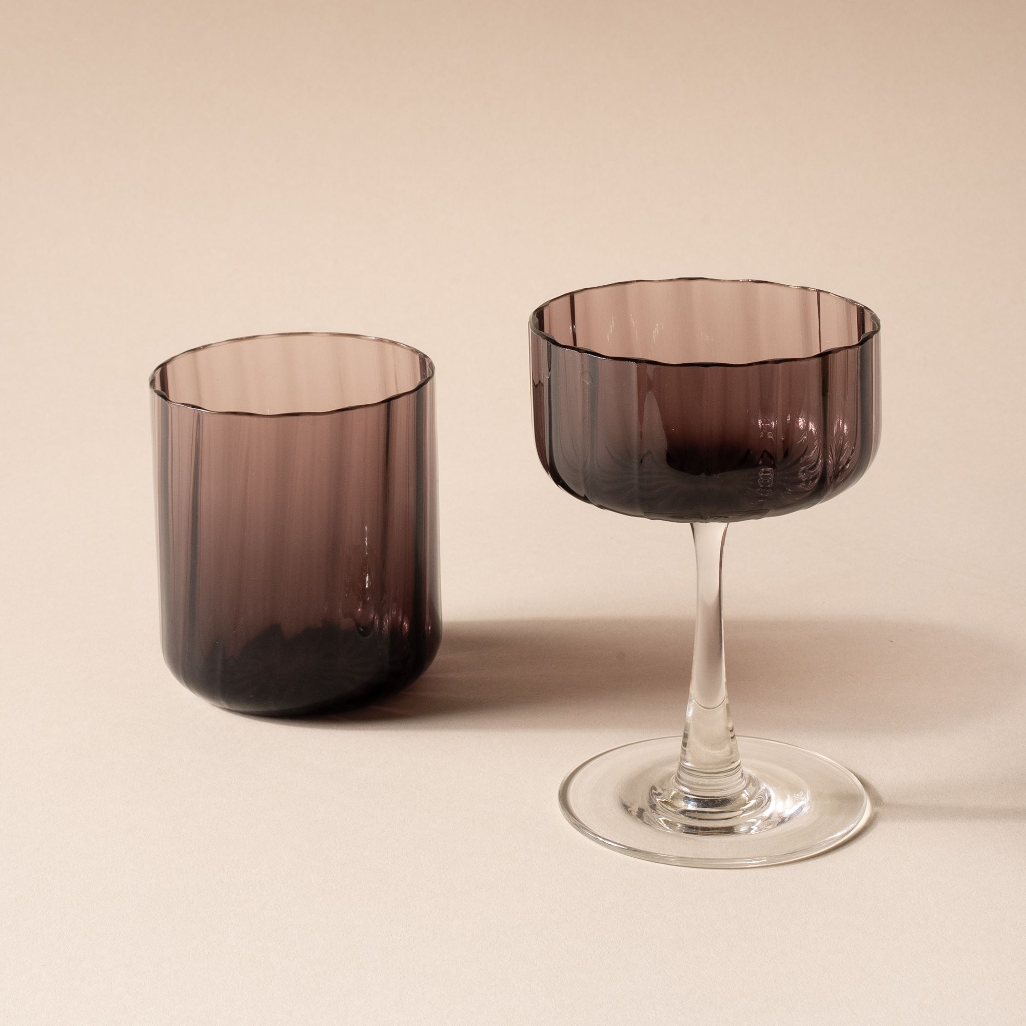 A fluted walled tumbler and coupe glass with a fluted bowl that is a grey purple color with a clear stem