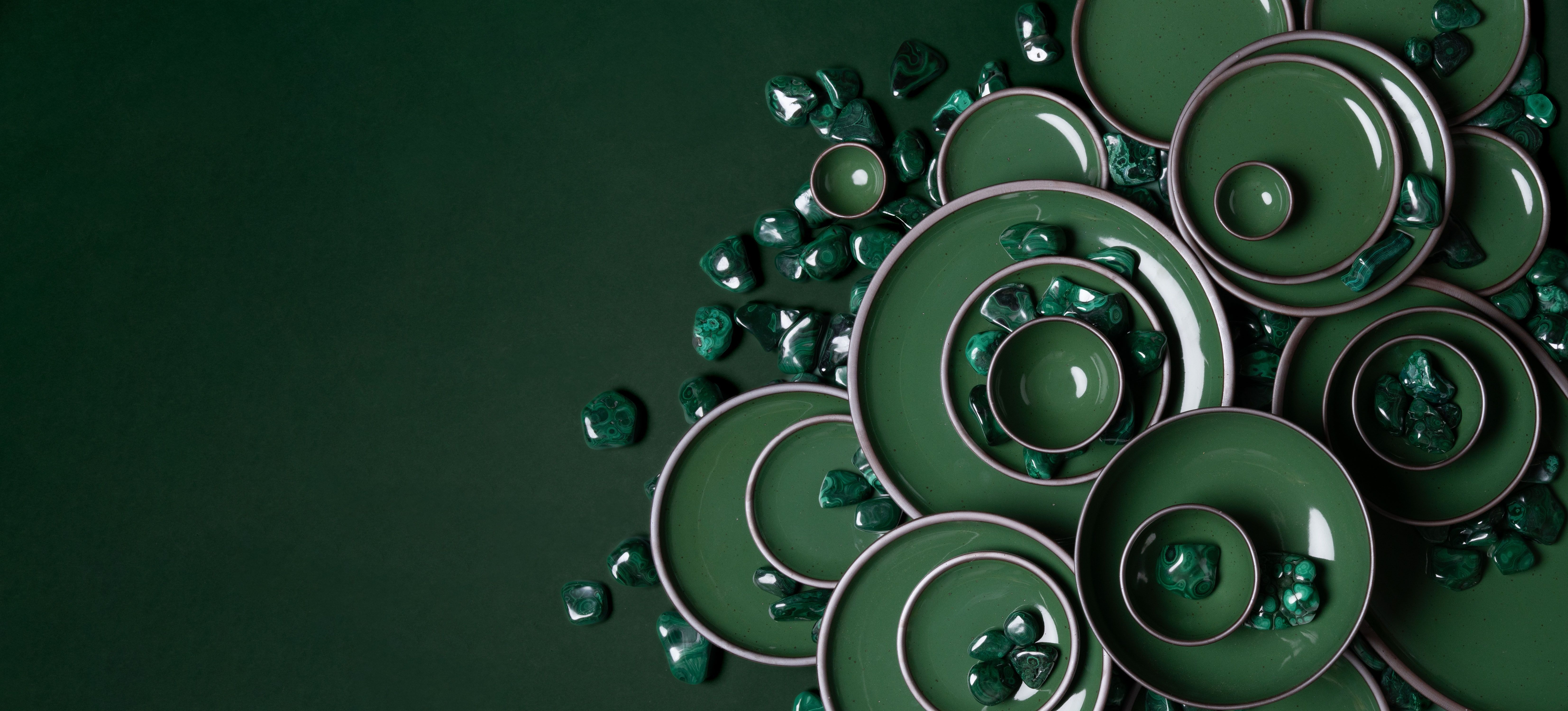 Arrangement of glossy emerald green ceramic plates and bowls with scattered malachite stones on a dark green background.