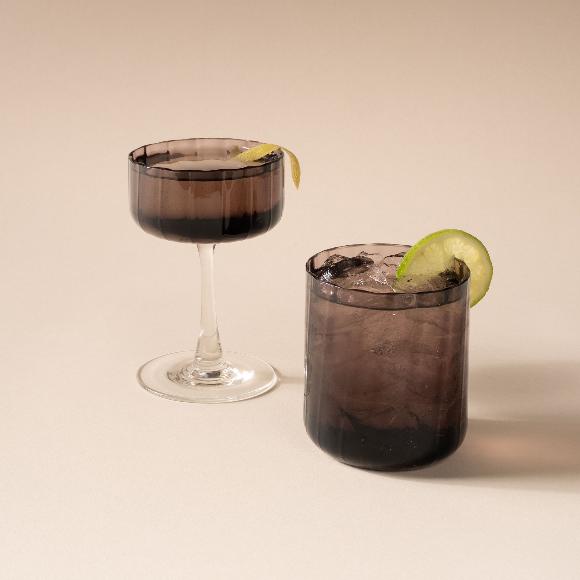 Cocktails in a fluted walled tumbler and coupe glass with a fluted bowl that is a grey purple color with a clear stem