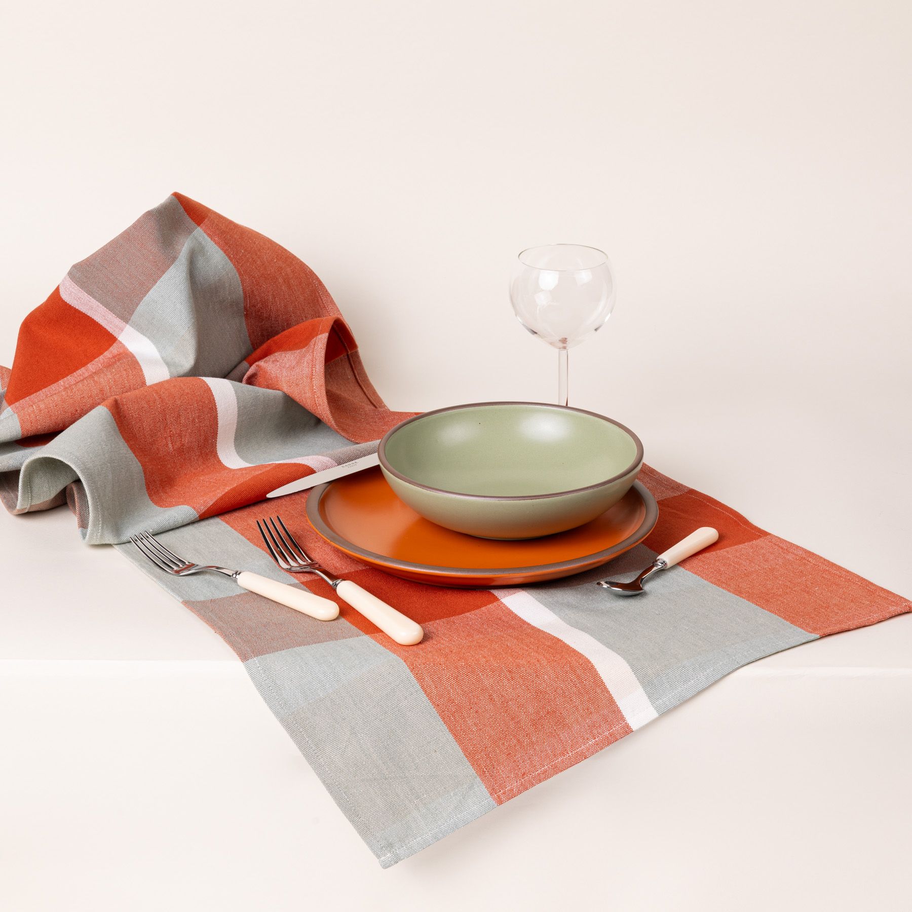 A gingham table runner featuring colors of sage green, bold orange, and cream, paired with a place setting with matching pottery and a wine glass.