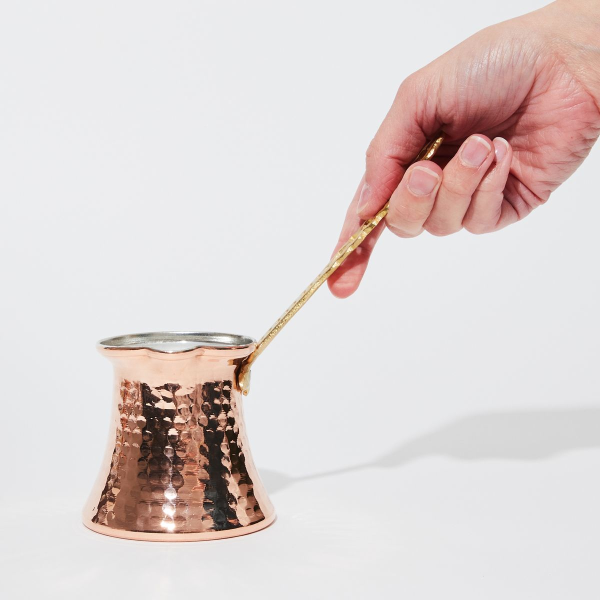 A hand holding a round copper pot by a long angled handle