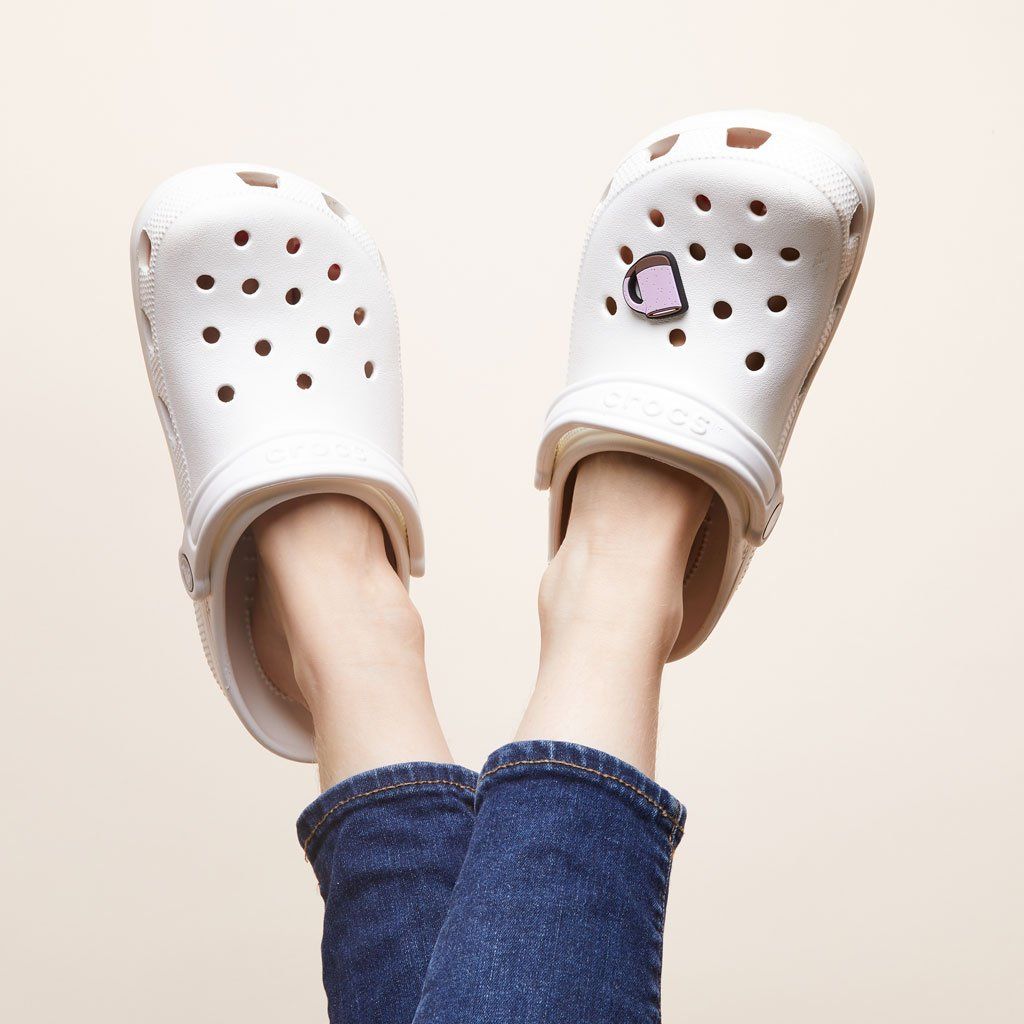 Two feet clad in white Crocs shoes. One has a charm that looks like a purple East Fork Mug. The model has bare ankles and medium blue jeans.