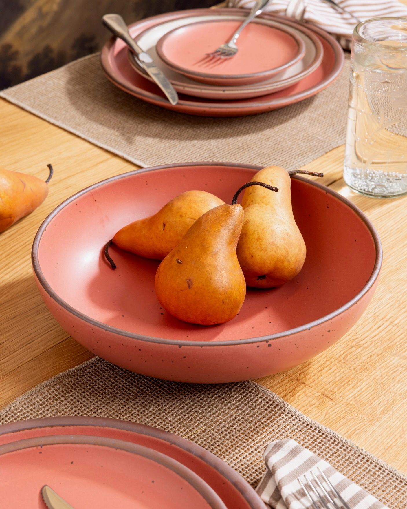 Guava-colored ceramic bowl filled with ripe pears on a wood table set with matching plates, glassware, and striped napkins.