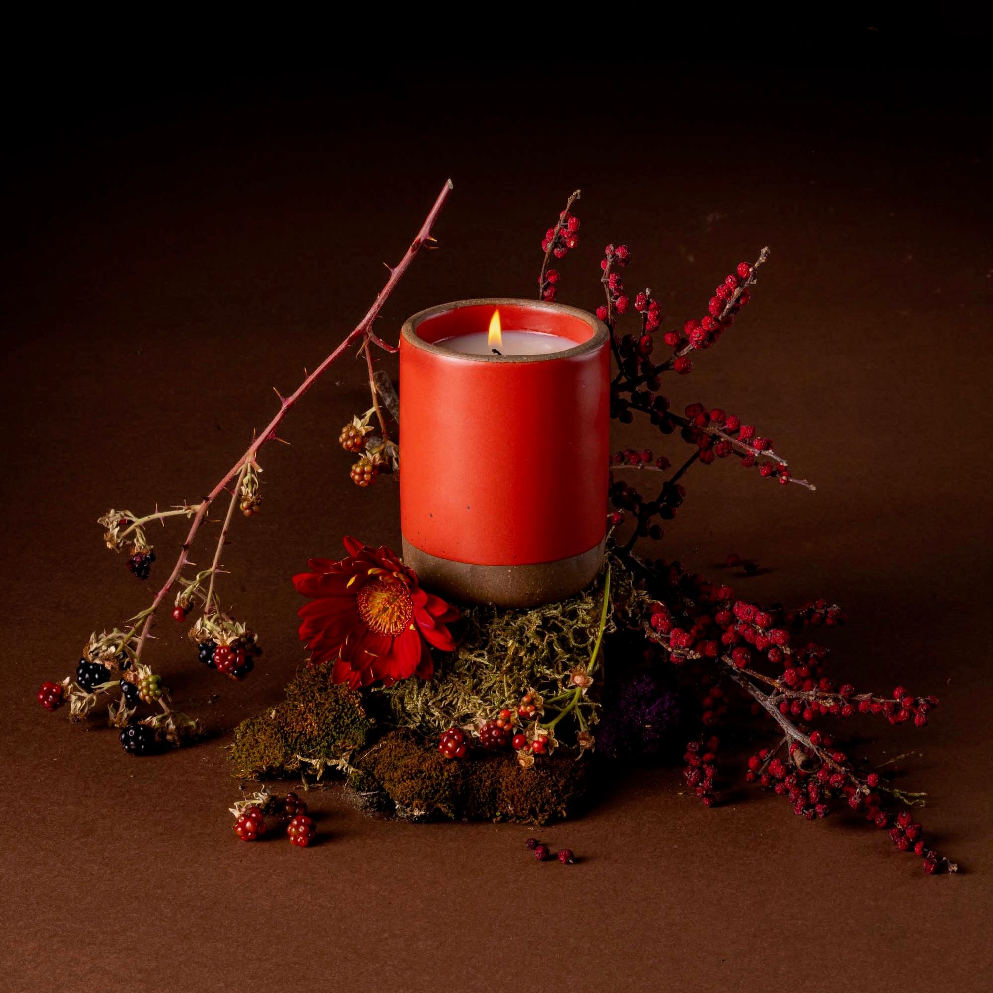 A bold red ceramic vessel with a lit candle inside sitting up on a small bed of moss and dirt, surrounded by berries and red branches with a brown background