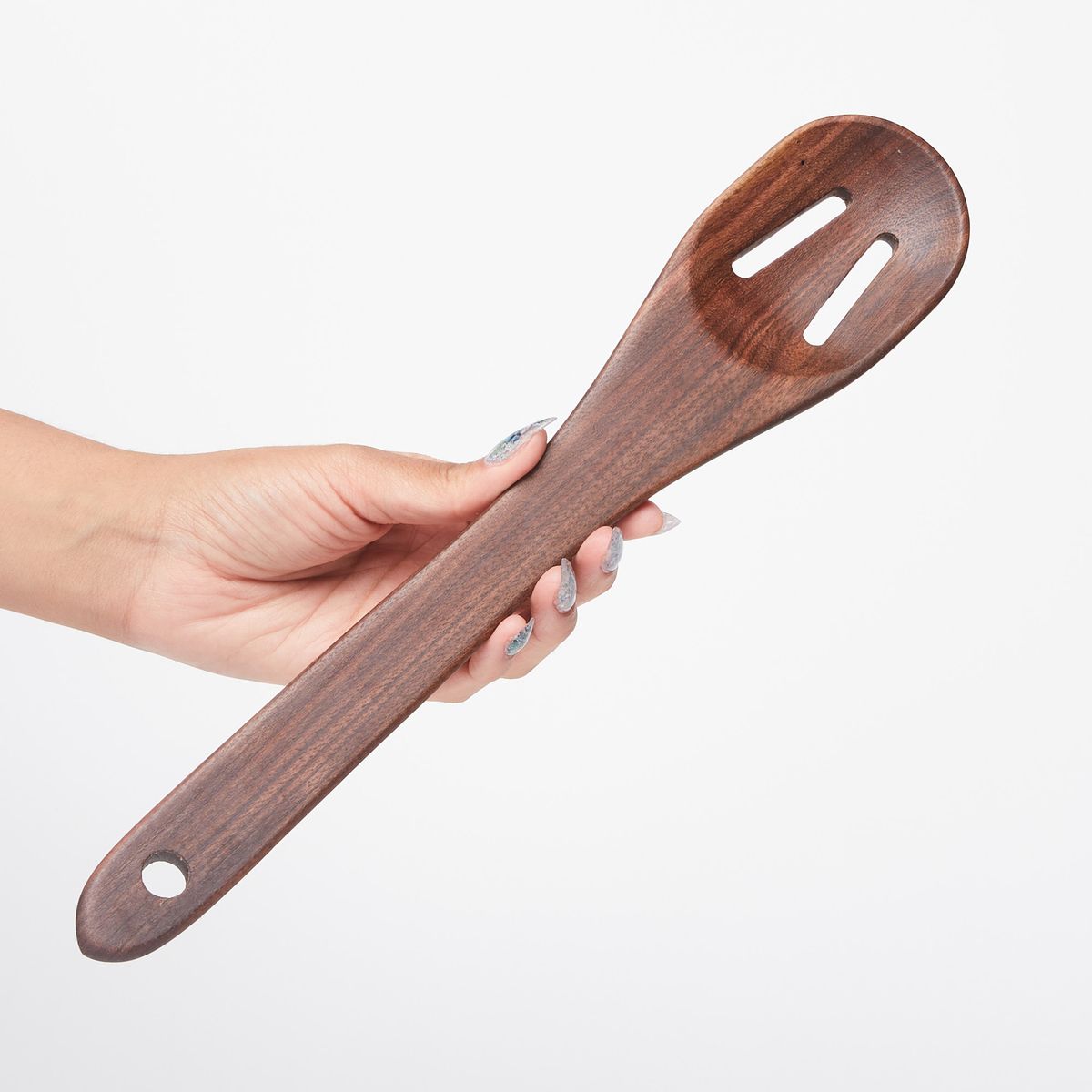 Hand holding a long black walnut wood spoon with two slots at the top and a single hole at the bottom