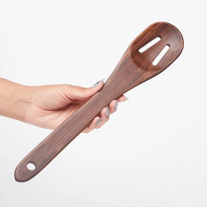Hand holding a long black walnut wood spoon with two slots at the top and a single hole at the bottom
