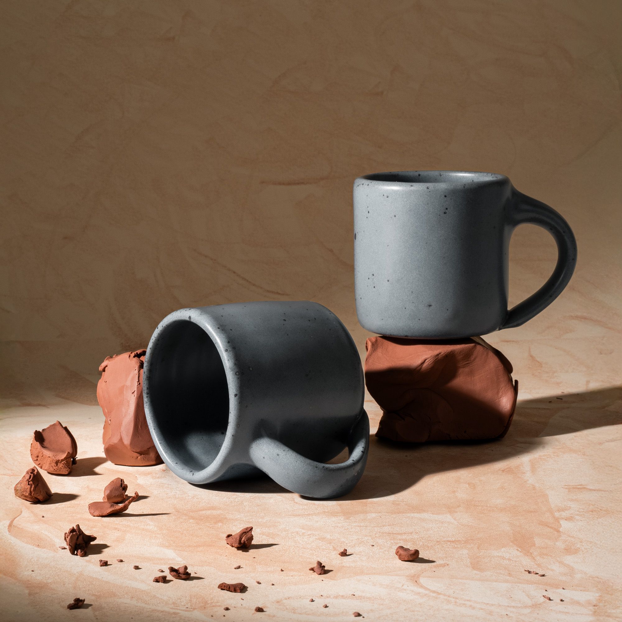 Two medium sized ceramic mugs with handles in a light blue-grey color featuring iron speckles, styled with raw clay