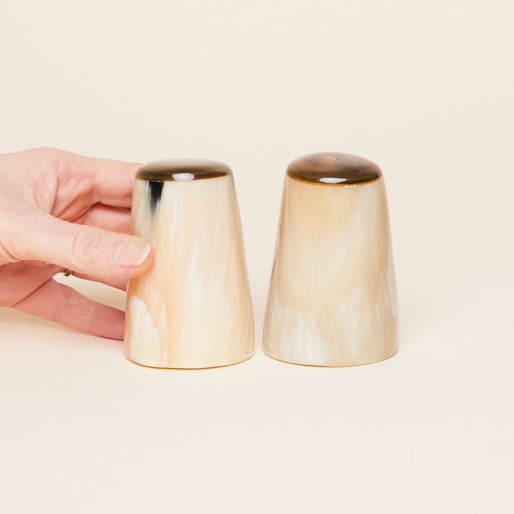 A hand touches one of two salt and pepper shakers that are cylindrical and flare toward the base in various shades of brown