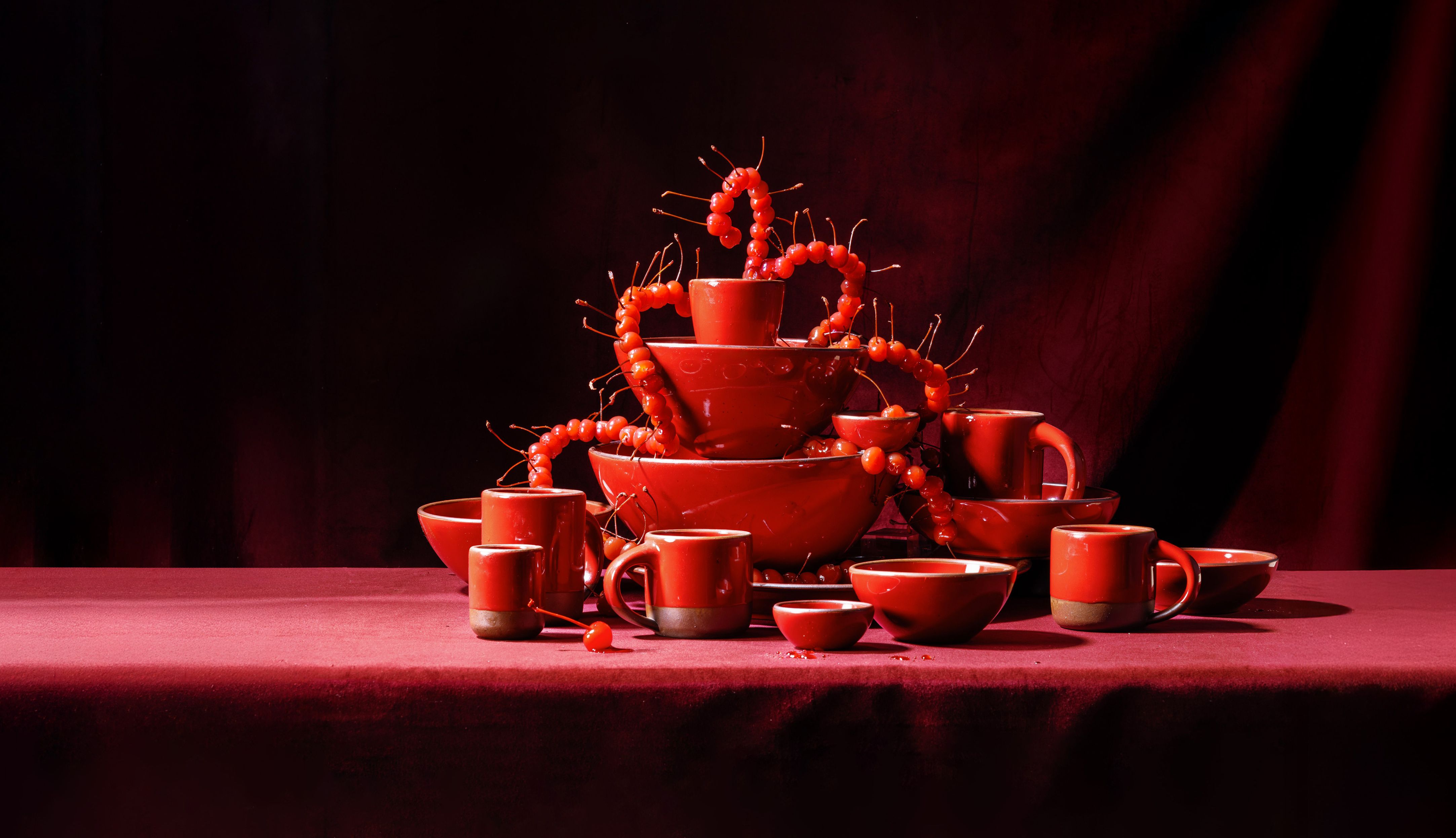Glossy red ceramic cups, mugs and bowls stacked into a tower, draped with glossy maraschino cherries against a deep red backdrop.